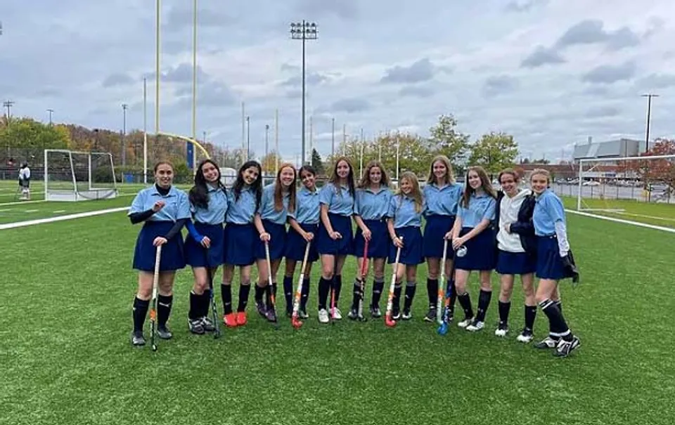Un gruppo di studentesse in divisa sportiva si trova sul campo da calcio della A.Y. Jackson Secondary School con il panorama urbano sullo sfondo.