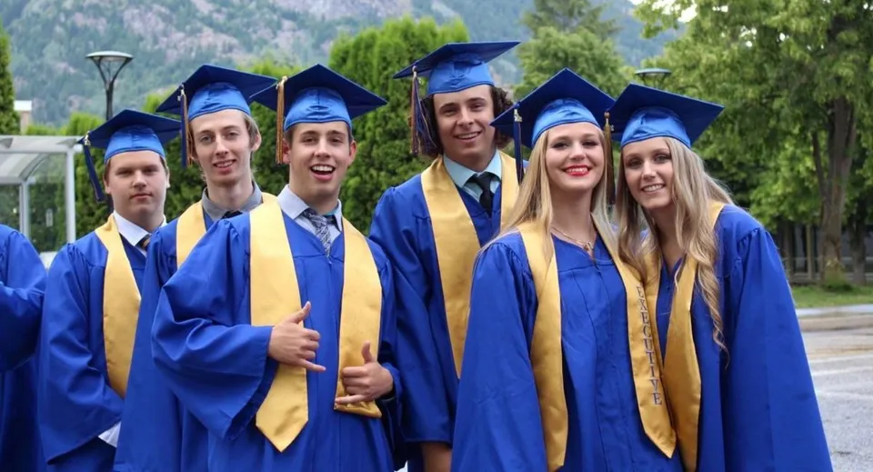 I diplomati della J.V. Humphries Secondary School posano insieme all'aperto con le loro toghe blu e dorate davanti ad alberi e montagne.