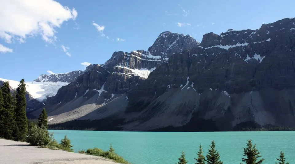 Un lago montano dalle acque turchesi davanti a una catena rocciosa parzialmente innevata con conifere.