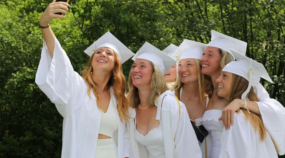 Un gruppo di diplomate della Proctor Academy è in piedi insieme all'aperto con toghe bianche da laurea.