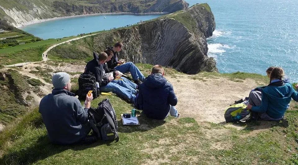 Studenti della Beech Grove School camminano su un sentiero erboso con vista su un paesaggio costiero roccioso e sul mare turchese.