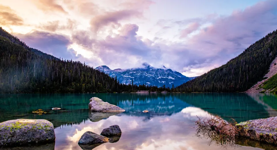 Un lago di montagna dalle acque turchesi con rocce nell'acqua, circondato da pendii boschivi e montagne tra le nuvole.