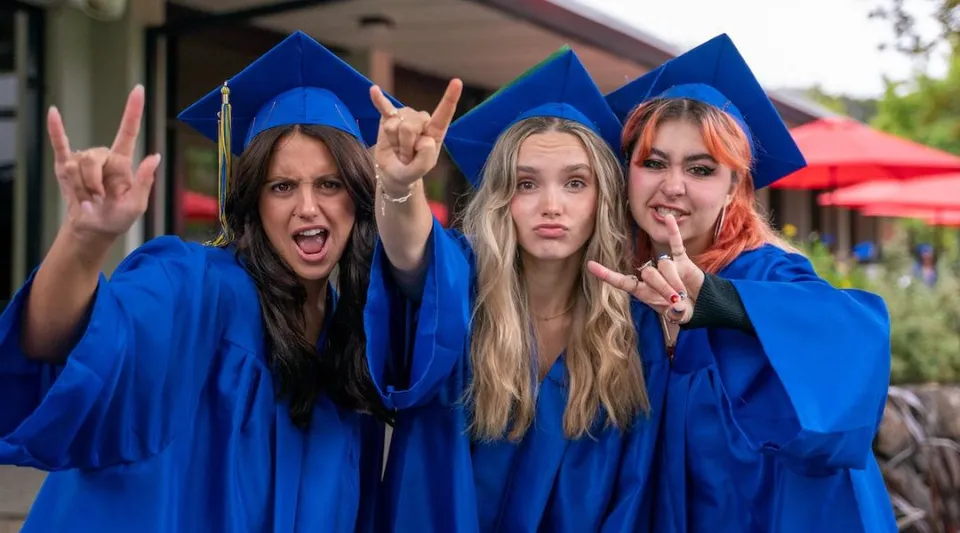 Tre studentesse della San Domenico School indossano toge blu per il diploma e posano davanti a una tenda rossa con il simbolo della pace.