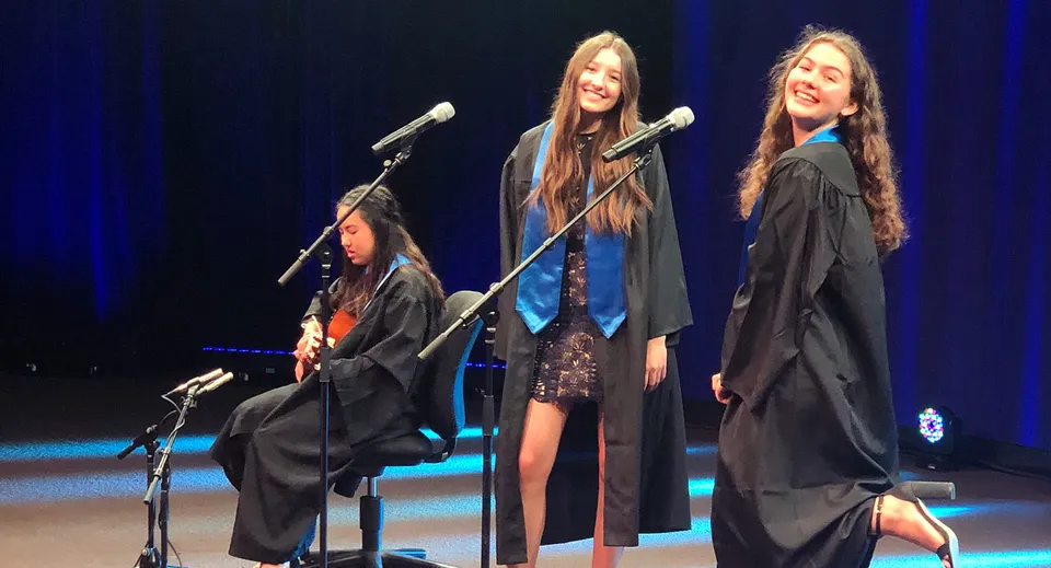 Tre studentesse della Alpha School sono sul palco in abito da cerimonia di diploma e parlano ai microfoni davanti a uno sfondo illuminato di blu.