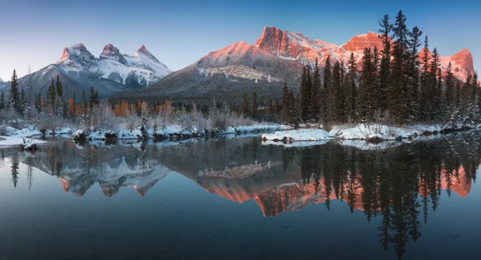 Montagne innevate e pini si riflettono nelle acque calme di un fiume nella natura canadese.
