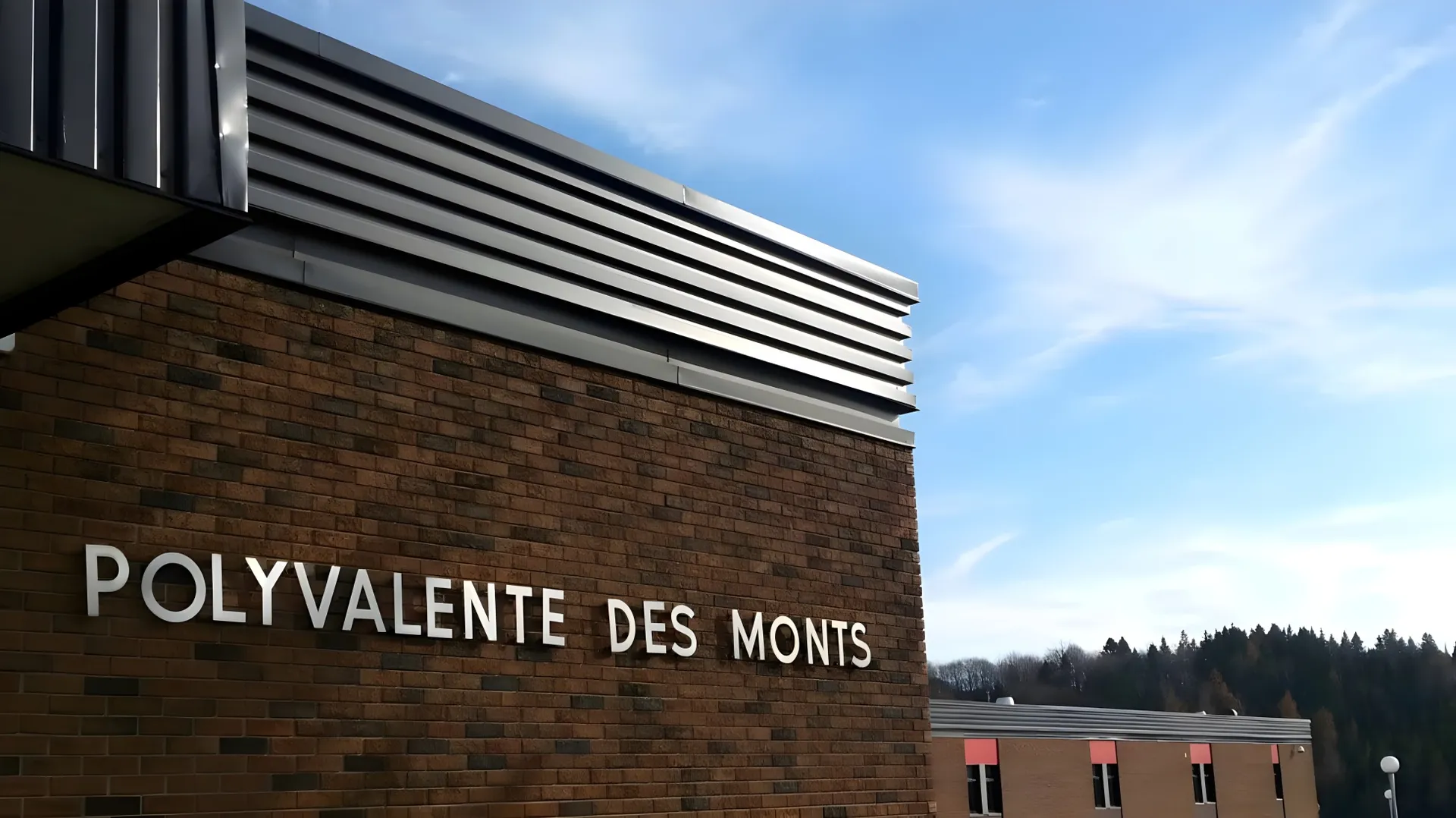 L'edificio moderno in mattoni della École Polyvalente des Monts si trova davanti a un cielo blu e colline boscose.