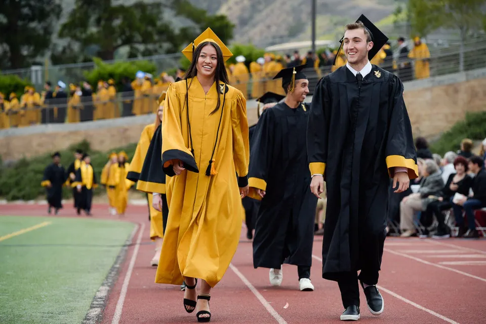 Due diplomati con toghe di colore diverso camminano sul campo sportivo dell'Oak Park Unified School District accanto a una folla di persone.