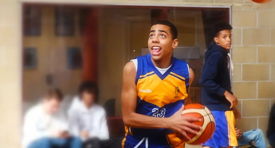 Uno studente del College of Richard Collyer tiene in mano un pallone da basket in una palestra con altre persone sullo sfondo.