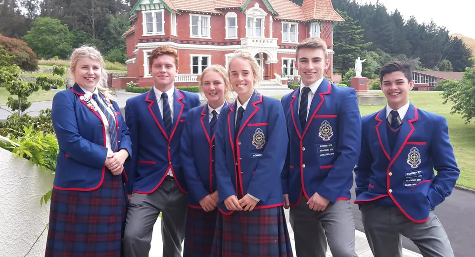Un gruppo di studenti del St. Kevin’s College in posa davanti a un grande edificio rosso e bianco circondato dal verde.