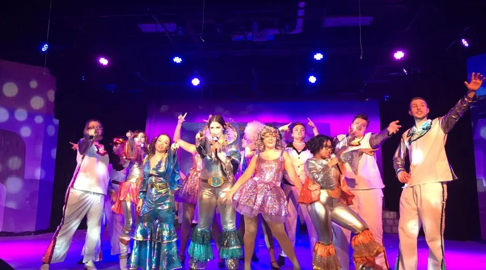 Studenti della Westwood Collegiate si esibiscono su un palco illuminato indossando costumi luccicanti durante uno spettacolo teatrale.