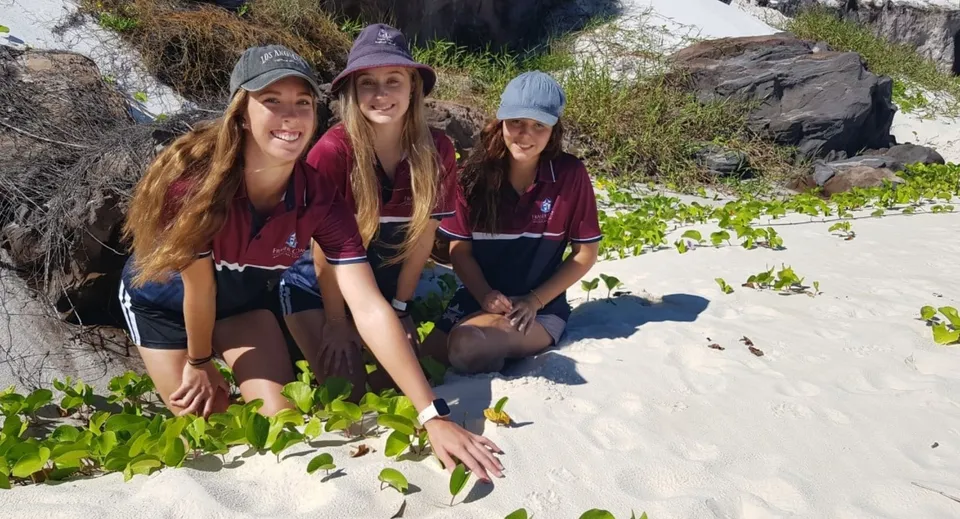 Tre studentesse del Fraser Coast Anglican College sono sulla spiaggia con sabbia, piante e rocce sullo sfondo.