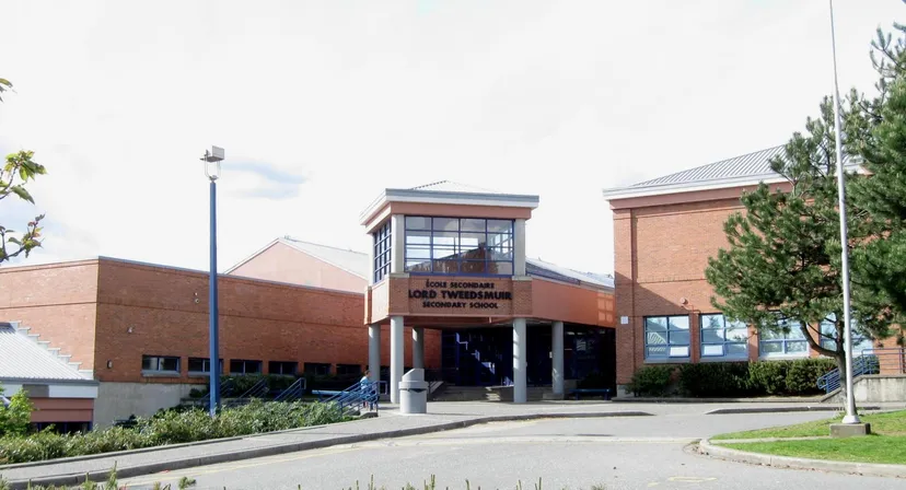 L'ampio edificio in mattoni con ingresso in vetro della Lord Tweedsmuir Secondary School è circondato da alberi e da un viale pavimentato.