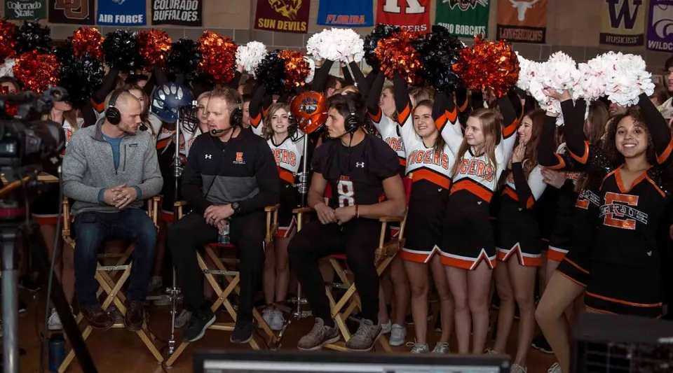 Un gruppo di cheerleader in divisa arancione e nera sta in piedi con i pompon davanti ai banner sportivi della Erie High School.