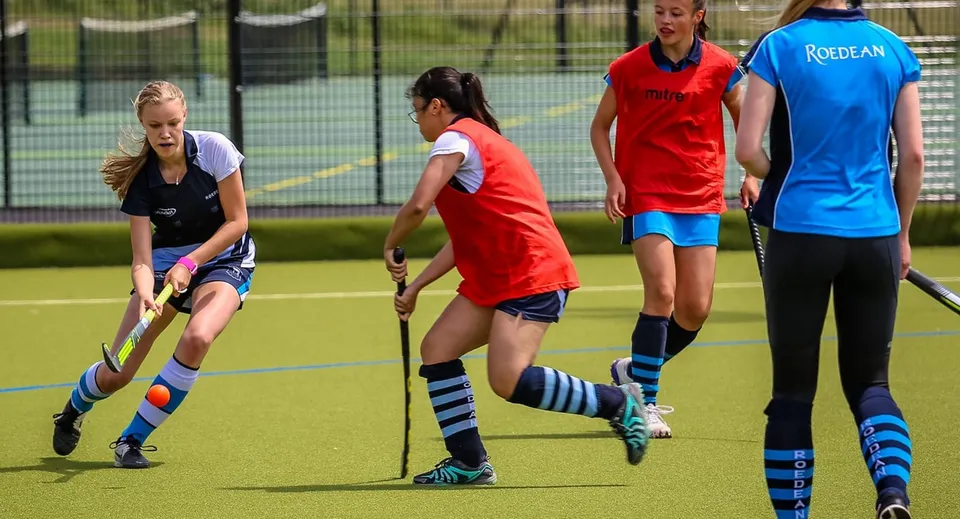 Studentesse della Roedean School giocano a hockey su prato su un campo in erba sintetica con una recinzione e alberi sullo sfondo.