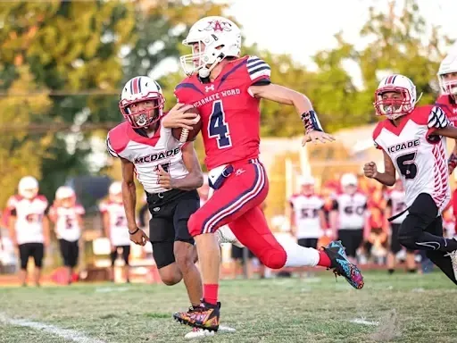 Estudiantes de la Incarnate Word Academy juegan al fútbol en el campo deportivo con espectadores en el fondo.