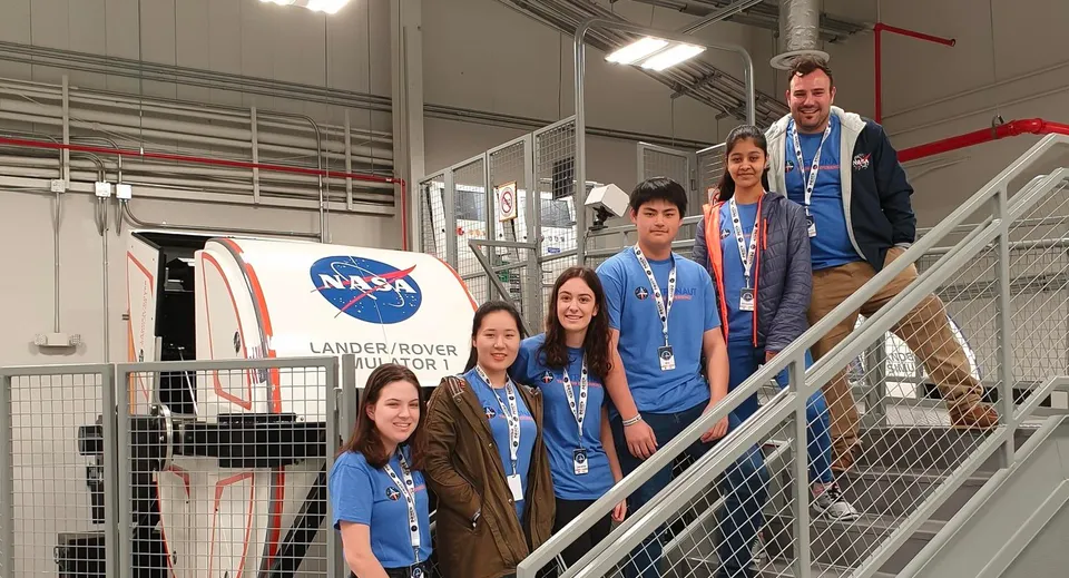Un gruppo di studenti della Norwood International High School in divisa NASA davanti a un grande logo NASA e a un modello di Lander Rover.