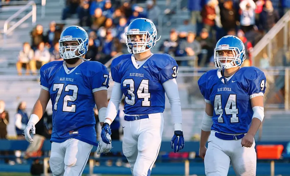 Tre giocatori di football in uniformi blu sul campo della Shore Regional High School davanti al pubblico.