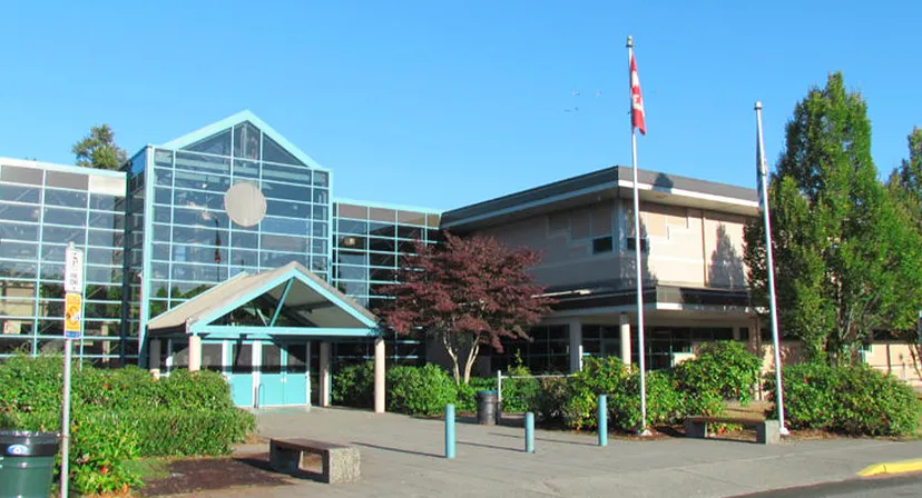 Si vede il moderno edificio in vetro e cemento della Walnut Grove Secondary School con la bandiera canadese in primo piano e circondato da alberi.