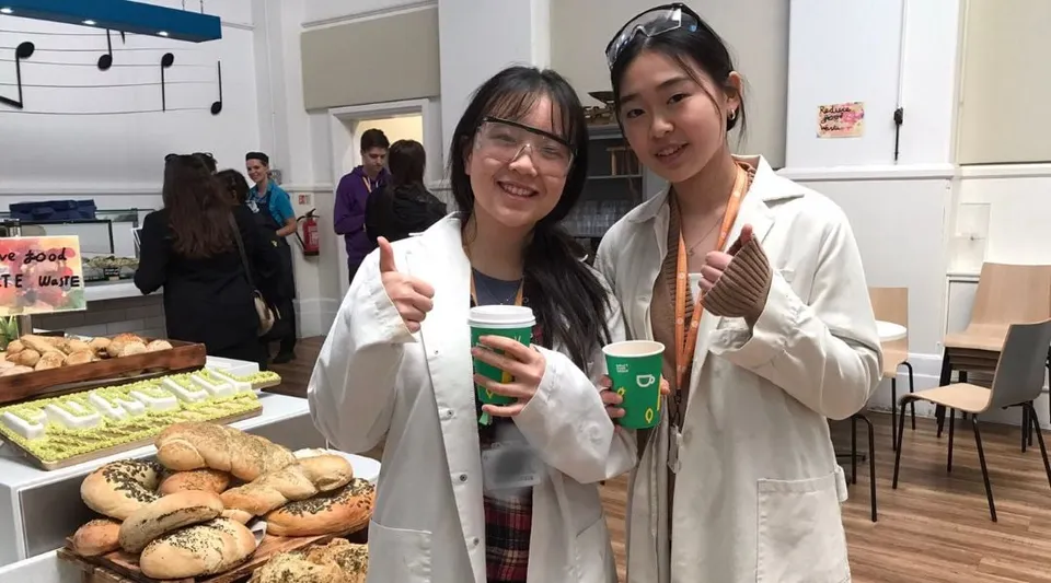Due studentesse della The Purcell School for Young Musicians indossano camici da laboratorio e tengono in mano tazzine da caffè in una stanza con prodotti da forno.