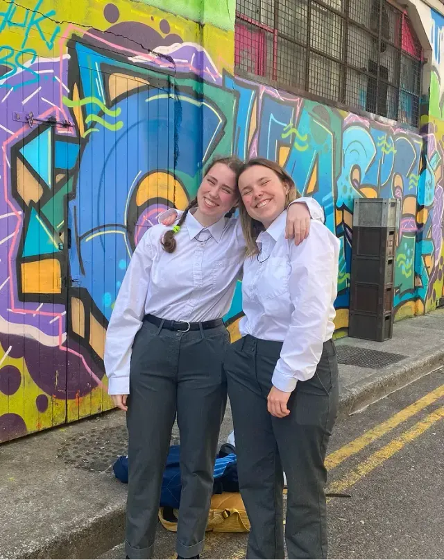 Due studentesse in uniforme scolastica sorridono davanti a un muro di graffiti