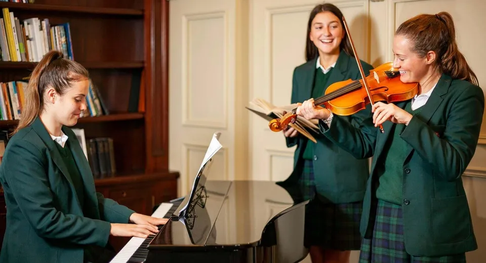 Tre studentesse della Salerno Secondary School suonano insieme il violino e il pianoforte in un'aula di musica con librerie.