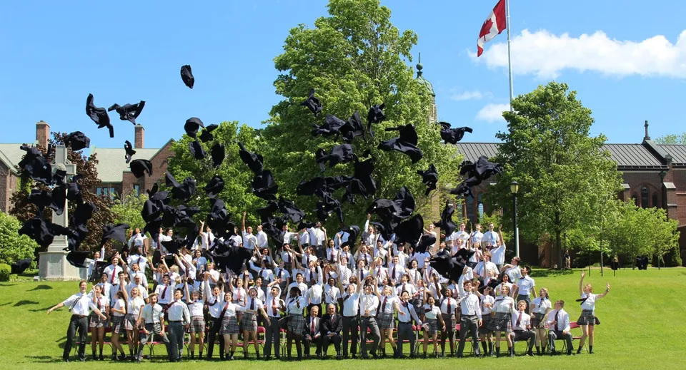 Un gruppo di diplomati e diplomate in toga bianca e tocco si riunisce sul prato della Trinity College School.