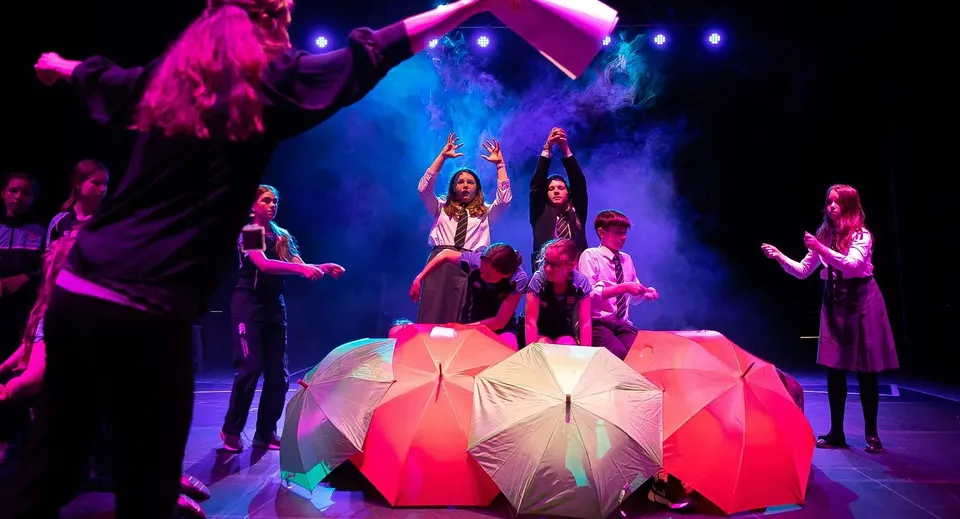 Gli studenti della Pocklington School mettono in scena uno spettacolo di danza con costumi colorati e ombrelli su un palco con un'illuminazione d'effetto.