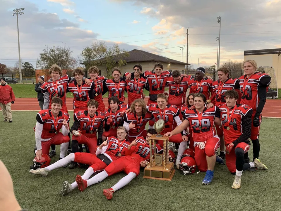 Un gruppo di giovani calciatori della Oakville Trafalgar High School posa con un trofeo su un campo sportivo.