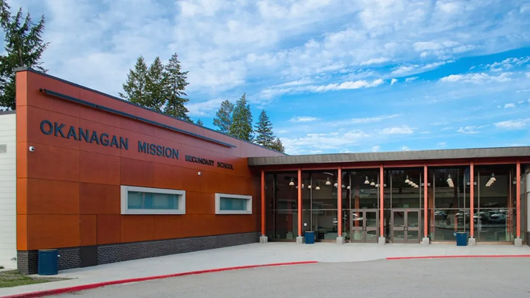 El moderno edificio naranja de la Okanagan Mission Secondary School con grandes ventanas y pinos en los alrededores.