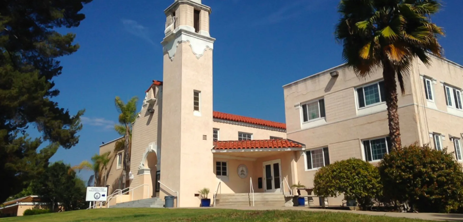 L'edificio di Christian Unified Schools con una torre, tegole rosse e una palma davanti a un cielo azzurro.