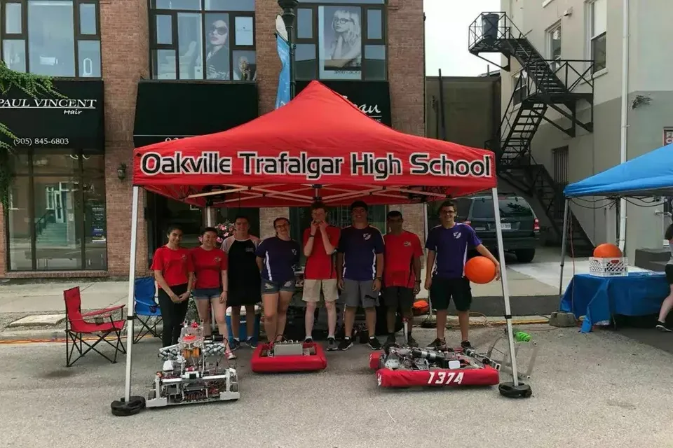 Un gruppo di studenti della Oakville Trafalgar High School sta davanti a una tenda rossa con il nome della scuola accanto a un robot e attrezzature a terra.