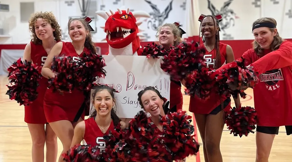 Un gruppo di cheerleader della Stuart Hall School posa in palestra con divise rosse e nere, pom-pom e un grande costume da mascotte rosso.