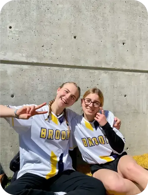 Dos estudiantes con camiseta deportiva hacen el signo de la paz sentadas contra un muro