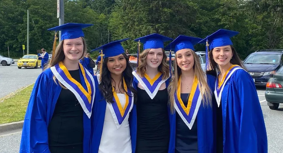 Cinque studentesse in abiti da diploma blu e gialli sono insieme all'aperto nel campus della Howe Sound Secondary School.