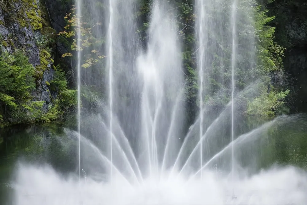 Große Wasserfontäne über einem Teich vor einer bemoosten Felswand mit Nadelbäumen