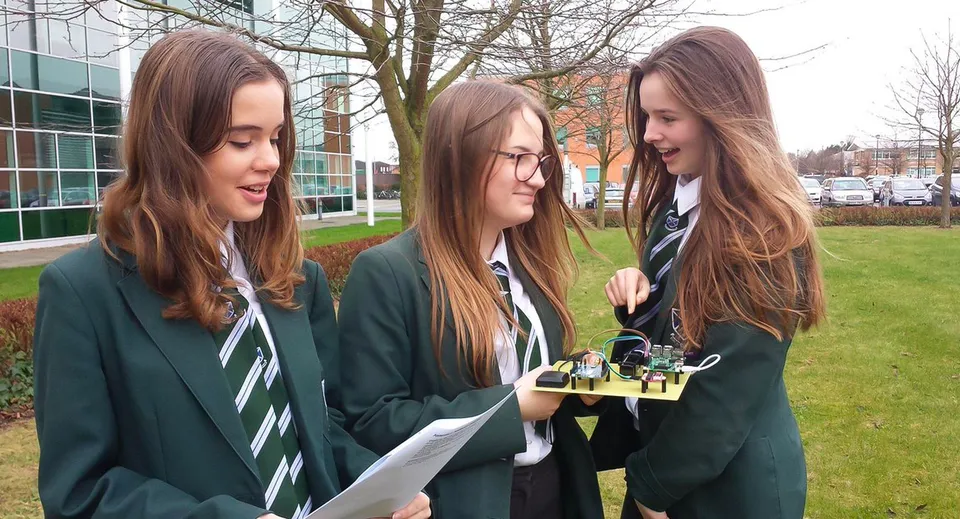 Tre studentesse in divisa scolastica si trovano nel campus della St. Alban's Catholic High School con l'edificio e gli alberi sullo sfondo.