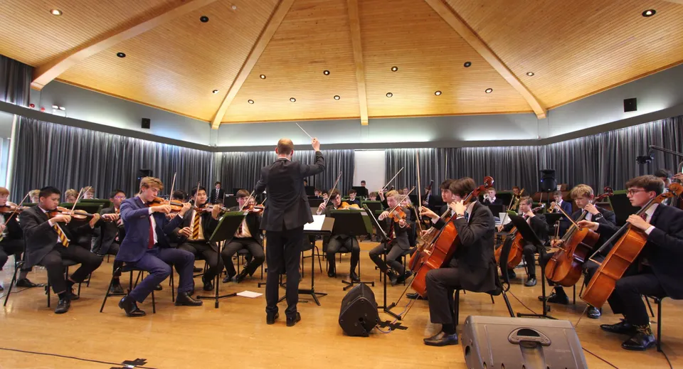 L'orchestra del Radley College esegue un concerto sotto la guida del direttore su un palco con soffitto a volta in legno.