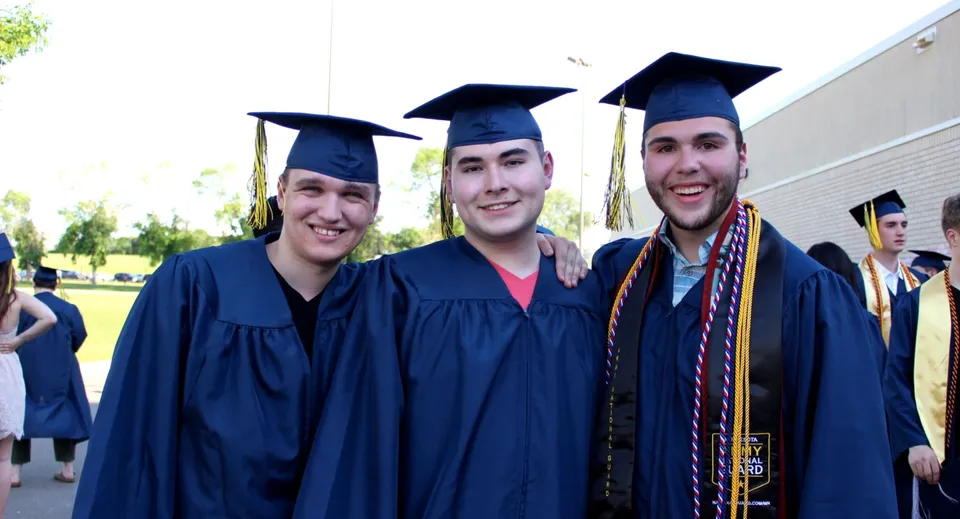 Tre studenti della Rosemount High School festeggiano il loro diploma in toga nell'area scolastica con spazi verdi sullo sfondo.