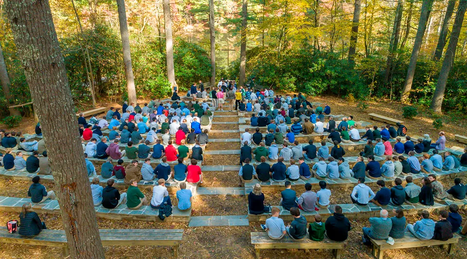Un grande gruppo di persone seduto su panche in legno nel bosco alla Christ School.