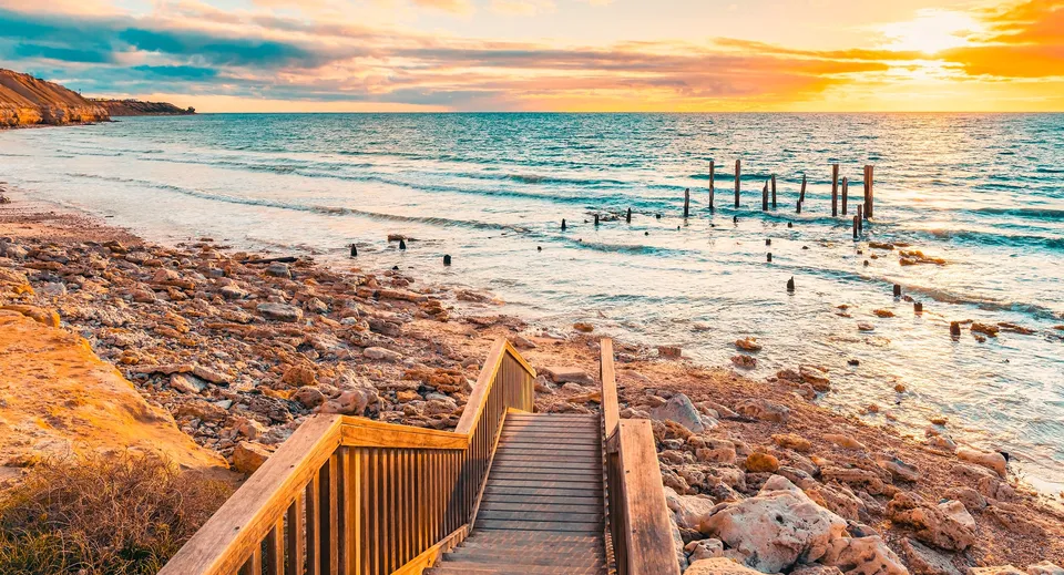 Una passerella in legno porta a una spiaggia rocciosa con acque calme sotto un tramonto colorato vicino alla Willunga High School.