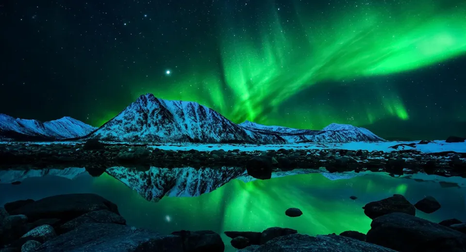 L'immagine mostra l'aurora boreale sopra un lago calmo di notte, con il riflesso delle montagne innevate.