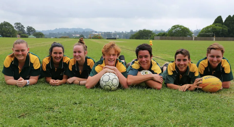 Un gruppo di giovani calciatrici siede su un campo da gioco erboso della Kenmore State High School, circondato da alberi e da un cielo nuvoloso.