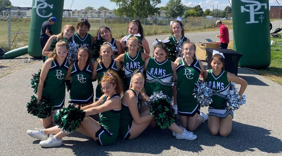 Un gruppo di cheerleader della Billings Central Catholic High School posa all'aperto davanti a un campo sportivo recintato.