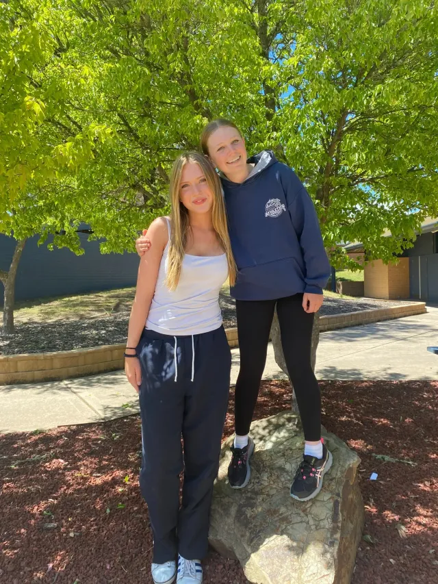 Due studentesse in felpa sportiva posano abbracciate sotto un albero nel cortile di una scuola australiana