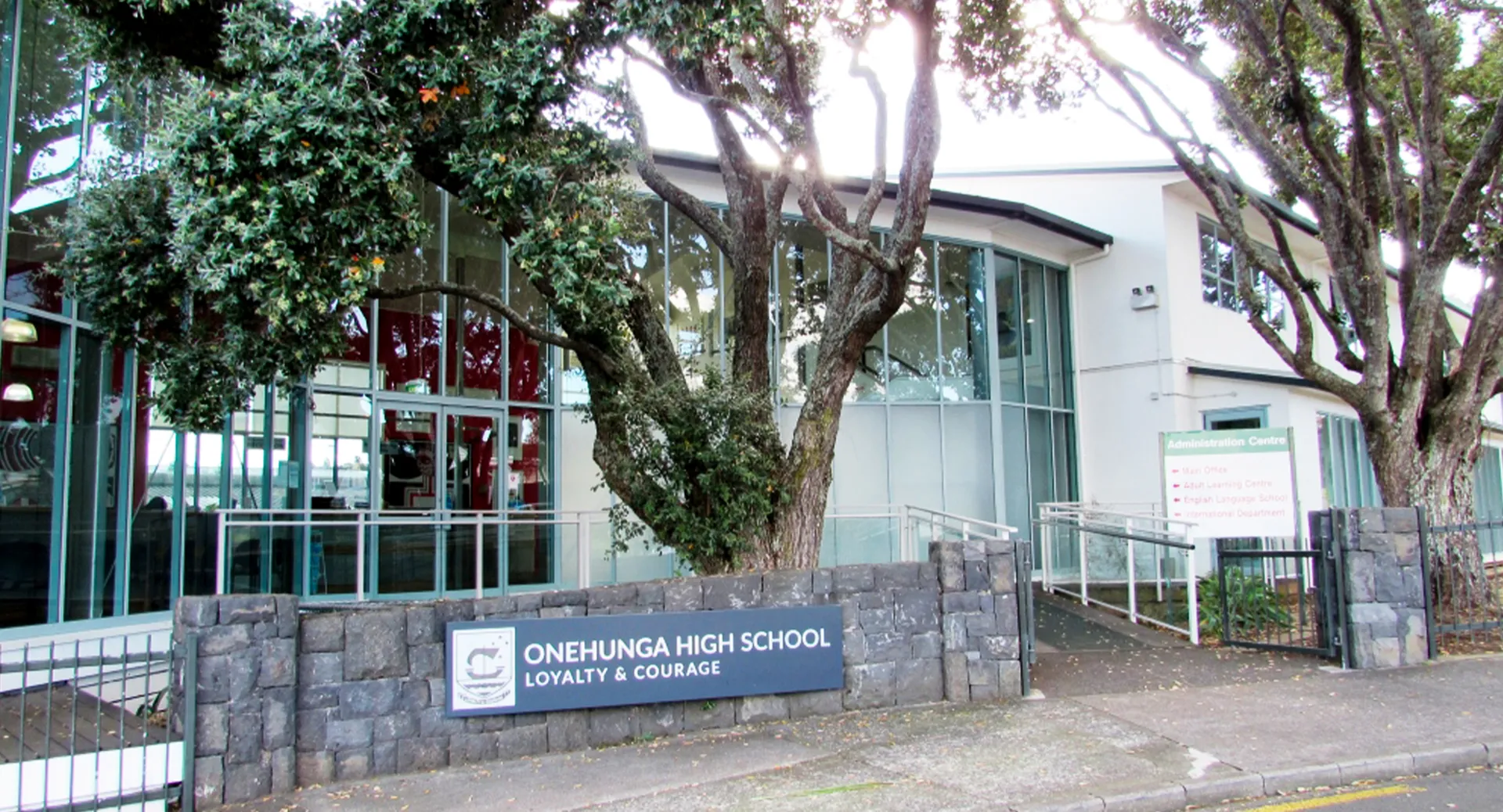 L'insegna in pietra della Onehunga High School si trova davanti a un edificio moderno, circondata da vegetazione verde.