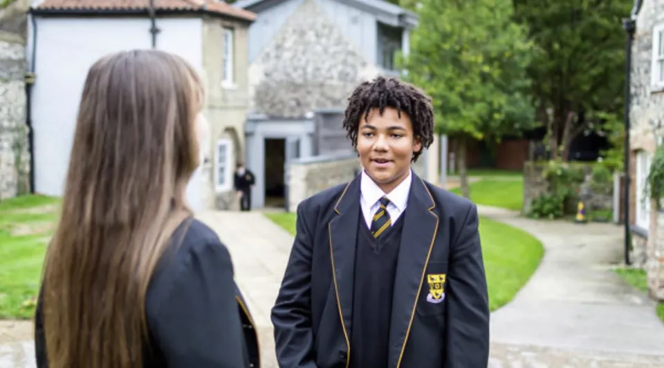 Due studenti camminano insieme lungo un viale tra aree verdi e un edificio sullo sfondo nel campus della Thetford Grammar School.