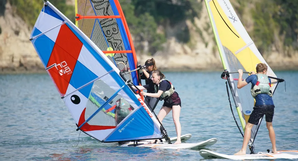 Barche a vela e windsurf navigano su uno specchio d'acqua calmo vicino al Wentworth College, circondati da un paesaggio boscoso.
