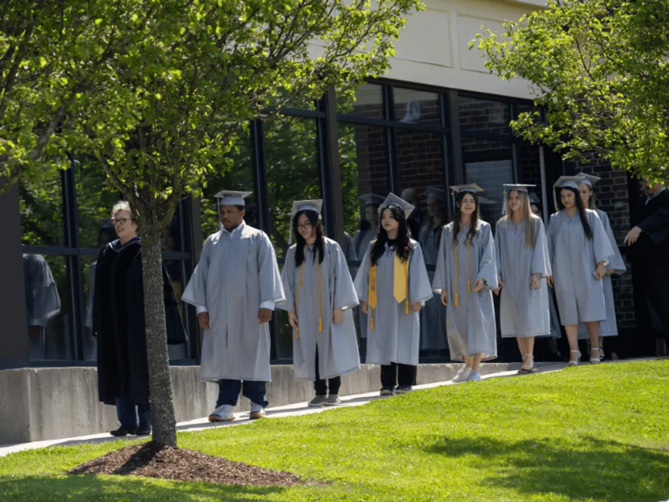 Studenti e studentesse diplomati della CATS Academy Boston in toga bianca su un prato davanti a un edificio.