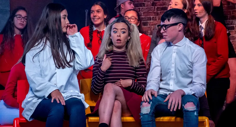 Un gruppo di studenti della St. Aidan's Comprehensive School è seduto sul palco davanti a una parete in mattoni durante una rappresentazione.
