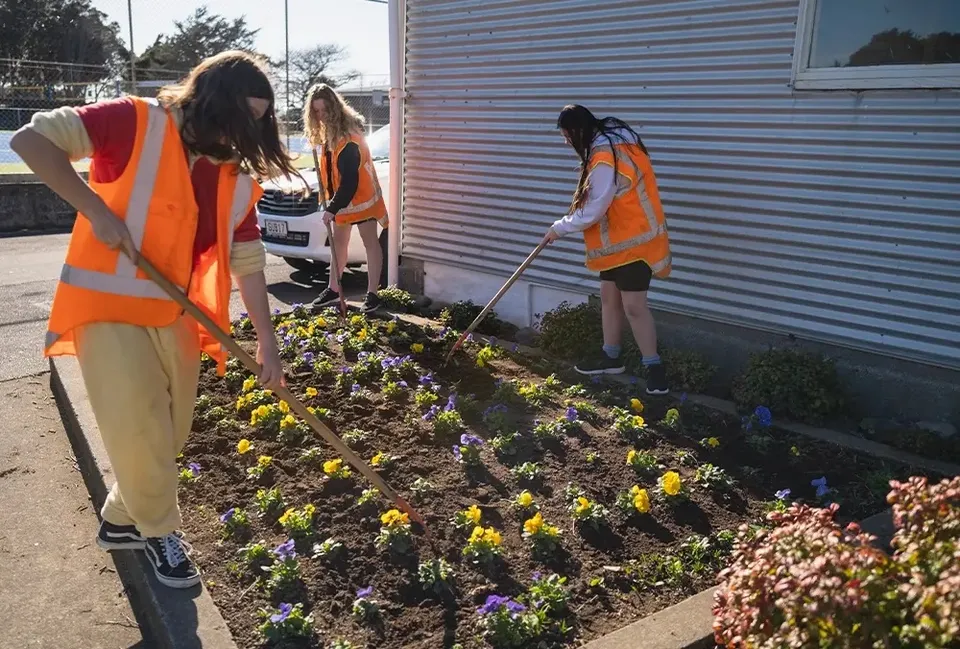 Studenti dello Spotswood College lavorano con gilet ad alta visibilità a un'aiuola davanti a una parete metallica.